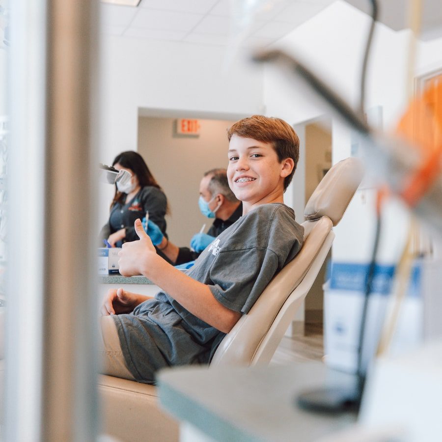 A boy sits in a dentist chair, smiling and giving a thumbs up