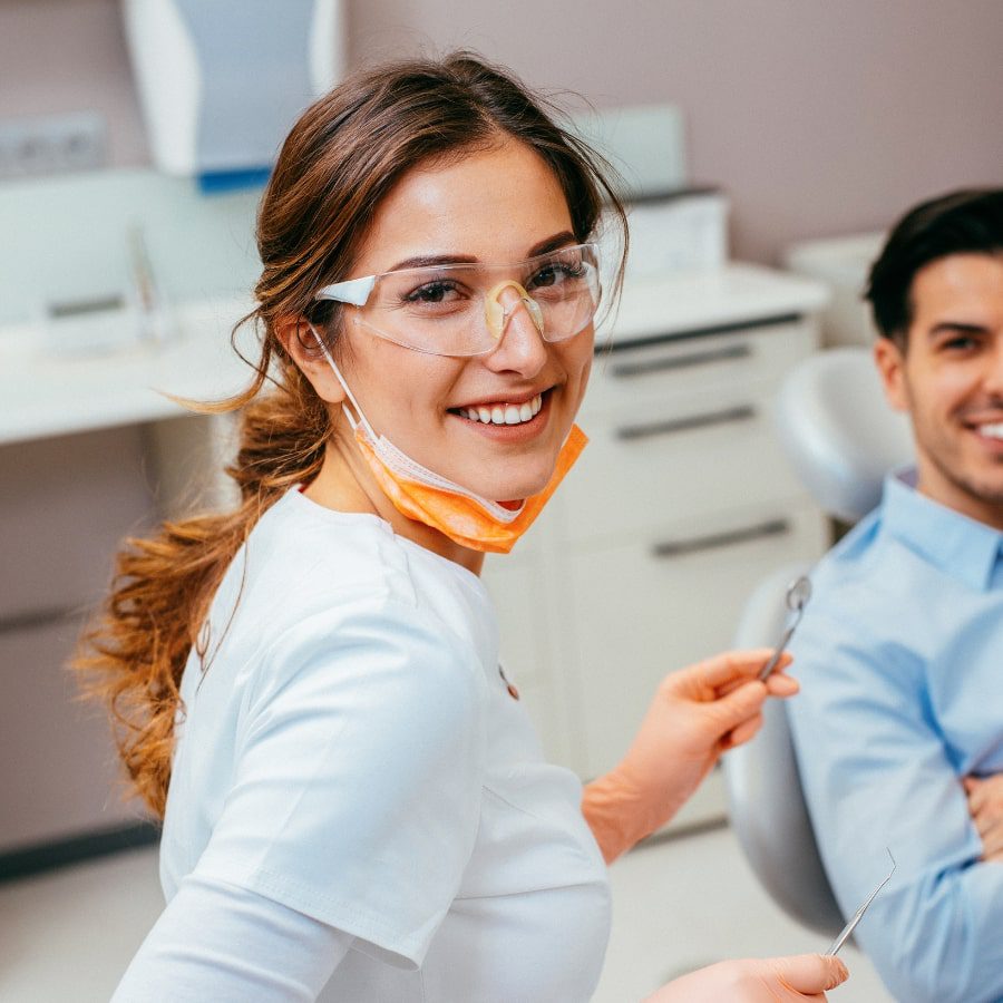 A dentist smiles at the camera, tools in hand and her patient in the background