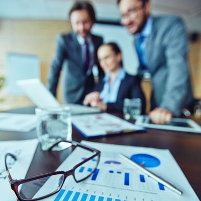 Three business professionals are gathered around a laptop, papers with graphs sit on the table in the foreground